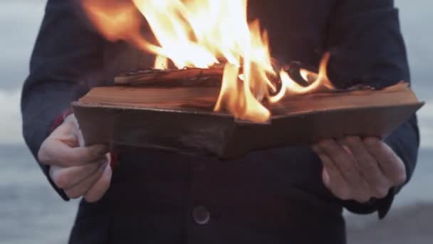 Giovane uomo con libro in fiamme in mano in piedi sulla costa nuvole tempesta sullo sfondo — Video Stock