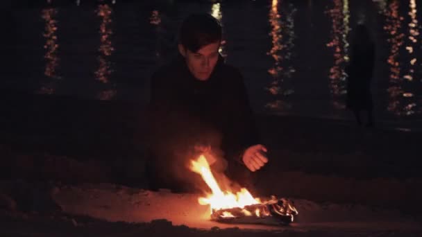 Jonge reiziger man zitplaatsen in de buurt vreugdevuur van brandende schoenen op het zand aan kust — Stockvideo