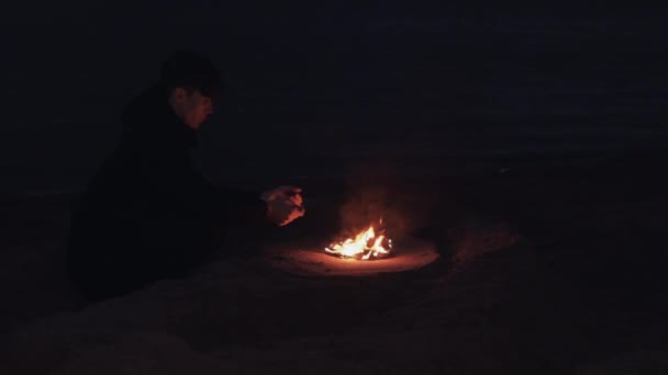 Jonge man zitten in de buurt vreugdevuur van brandende schoenen op het zand aan kust — Stockvideo