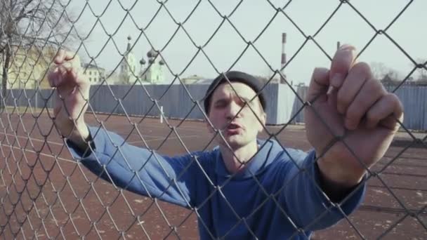 Agresivo caucásico hombre hablando algo en un campo de deportes detrás de valla de metal — Vídeos de Stock