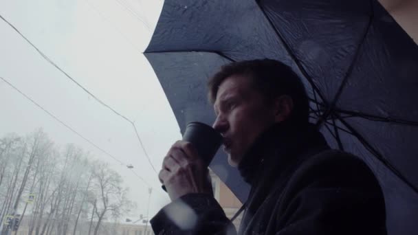 Jonge man met paraplu staan onder sneeuwval koffie drinken uit plastic beker — Stockvideo