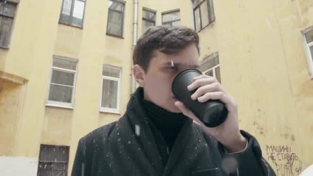 Jonge man met een vacht reputatie onder sneeuwval koffie drinken uit zwarte plastic beker — Stockvideo