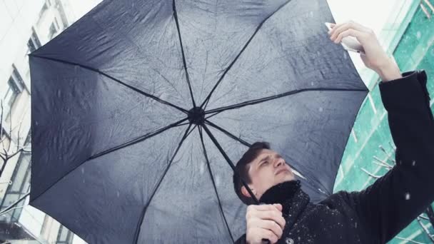 Молодой человек с зонтиком под снегопадом делает селфи фото с помощью смартфона — стоковое видео