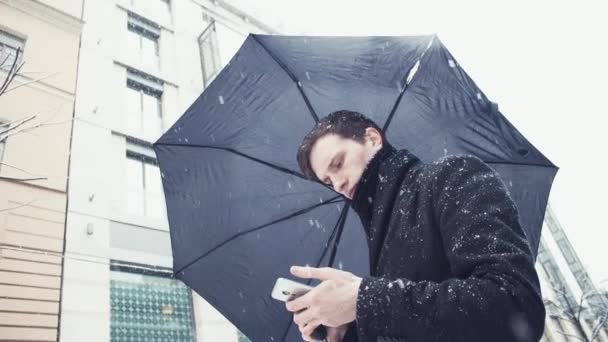 Mladý muž v plášti s deštníkem za sněžení pomocí smartphone na alej — Stock video