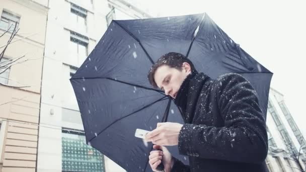 Young man in coat with umbrella under snowfall using smartphone on alley — Stock Video