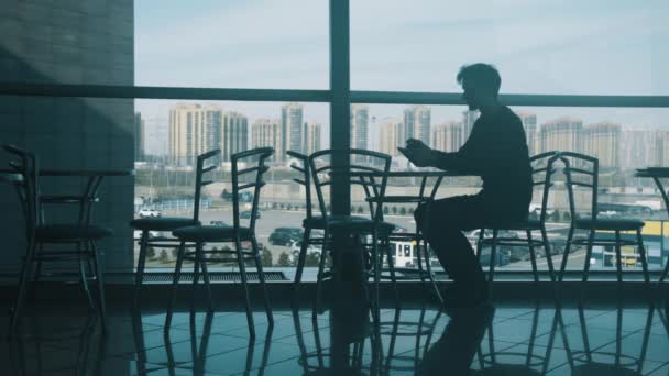 Hombre joven sentado solo en la mesa en los centros comerciales café con un teléfono inteligente — Vídeo de stock