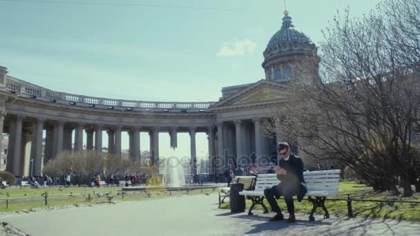 Ein junger Mann mit Smartphone sitzt auf einer Bank in der Nähe der orthodoxen Kirche. sonniger Tag — Stockvideo