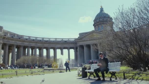 Молодой человек, пользующийся смартфоном, сидит на скамейке возле исторического здания. Солнечный день — стоковое видео