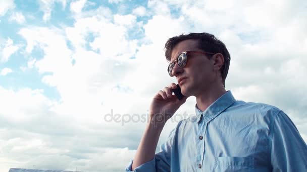 Young man talking on phone standing outdoors. Bright sky at background. — Stock Video