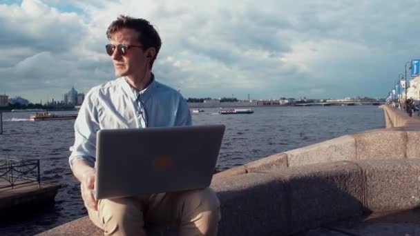 Молодий чоловік розмовляє на відеозв'язку з ноутбуком, що сидить на лавці біля набережної — стокове відео
