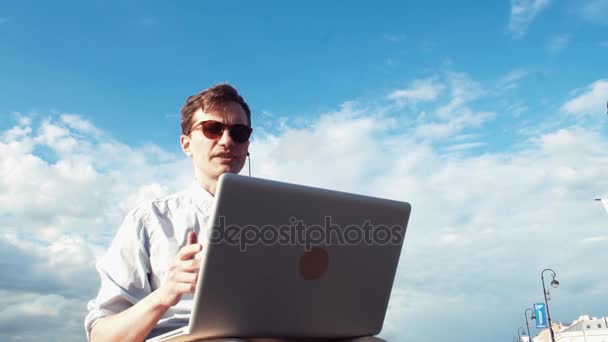 Νεαρός άνδρας έχοντας τηλεοπτική συνομιλία με το laptop κάθεται σε παγκάκι σε εξωτερικούς χώρους. Μέρα του καλοκαιριού — Αρχείο Βίντεο