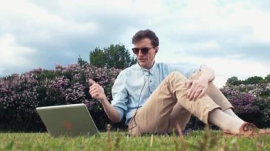 Genç adam Park yeşil çimenlerin üzerinde oturan laptop görüntülü sohbet sahip. Yaz günü
