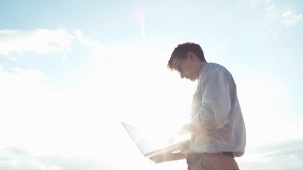 Jonge man vieren succes staande met laptop buiten. Felle zon schijnt. — Stockvideo