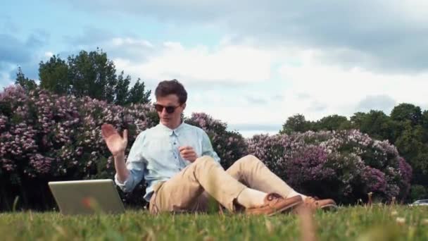 Фрилансер получает видеозвонок с ноутбука, сидящего на траве в городском парке. Летний день — стоковое видео
