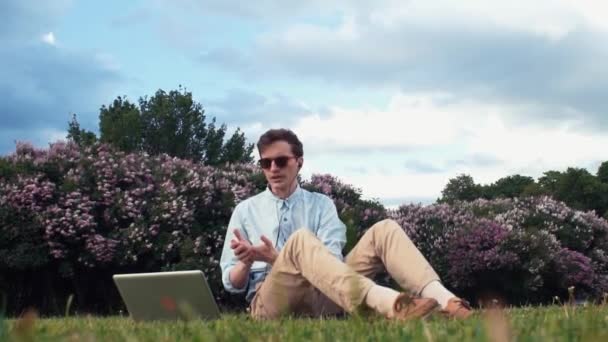 Young man having video call at laptop sitting on green grass at park. Summer day — Stock Video