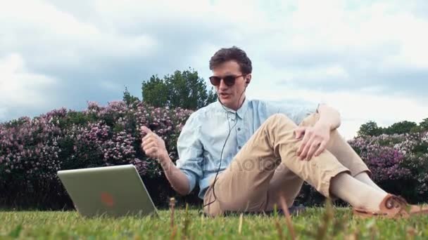 Young man having video chat at laptop sitting on green grass at park. Summer day — Stock Video