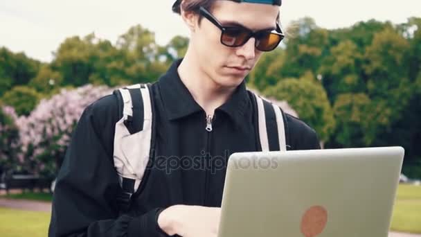 Poważny młody człowiek w okularach słonecznych i sprawny za pomocą laptopa stojący w parku — Wideo stockowe