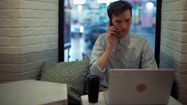 Młody mężczyzna rozmowa na telefon i za pomocą laptopa siedząc w kawiarni. Freelancer pracuje — Wideo stockowe