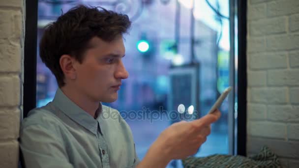 Caucásico joven escribiendo mensajes en el teléfono inteligente en la cafetería por la noche — Vídeo de stock