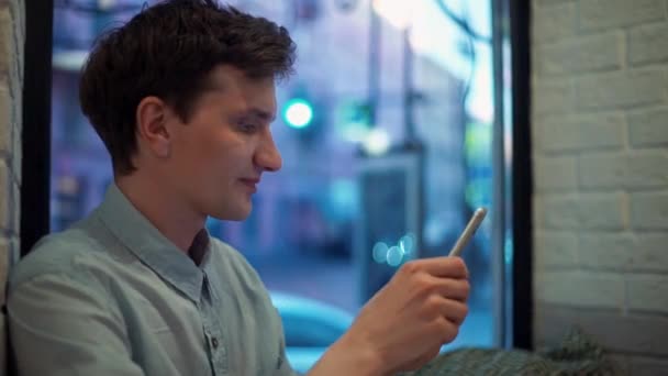 Sorrindo jovem digitando mensagens no smartphone no café à noite — Vídeo de Stock