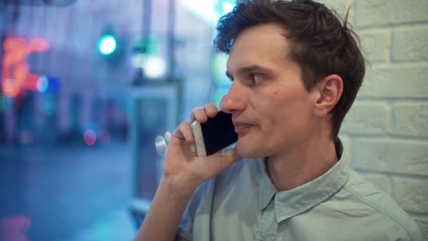 Joven con camisa azul hablando por teléfono en la cafetería por la noche — Vídeos de Stock
