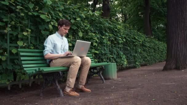 Jovem usando laptop com fones de ouvido sentado no banco no parque da cidade. Dia de verão — Vídeo de Stock