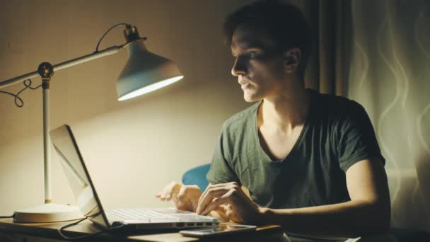 Jovem vestido com uma camiseta usando laptop em casa à noite — Vídeo de Stock