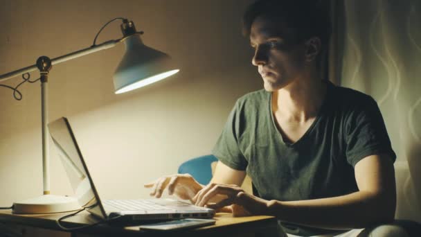 Young man dressed in a T-shirt using laptop at home in the evening — Stock Video