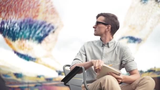 Young man sitting on chair at city park with a pencil and notebook in his hands — Stock Video