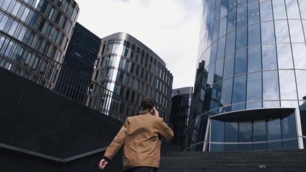 Молодой человек в дождевой куртке поднимается по лестнице в бизнес-центр, разговаривает по телефону — стоковое видео