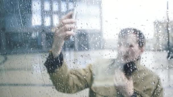 Homem de casaco usando smartphone sob chuva. Vista através da janela com gotas de chuva — Vídeo de Stock