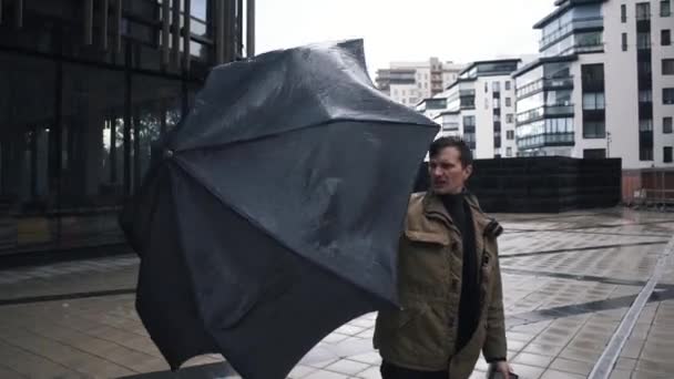 Молодой человек в куртке с зонтиком стоит возле здания бизнес-центра — стоковое видео