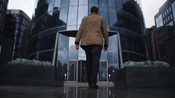Молодой бизнесмен в дождевой куртке идет к зданию бизнес-центра — стоковое видео