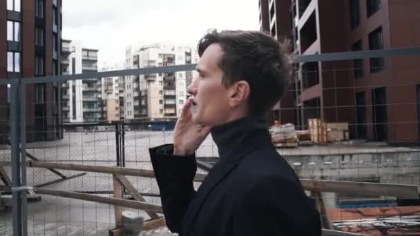 Junger Mann im Anzug mit Smartphone. Haus im Bau im Hintergrund — Stockvideo