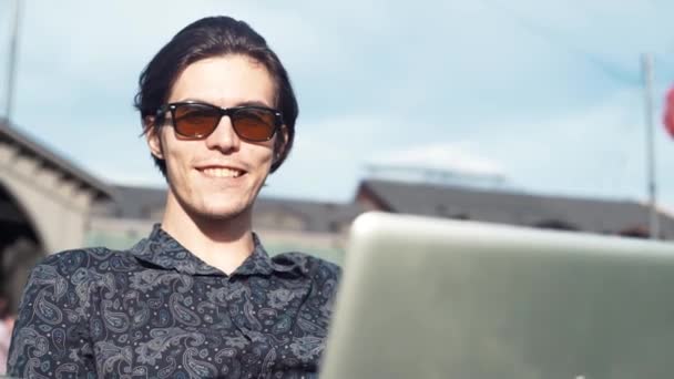 Fiatal férfi mosolyog nagyjából használ laptop ül a székre, a városi park. Nyári napon — Stock videók