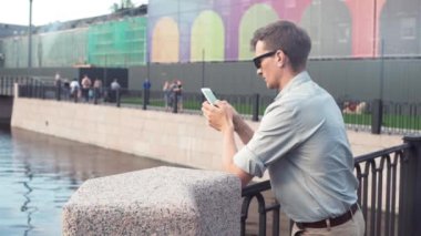 Smartphone ayakta şehir parkında kullanarak güneş gözlüklü genç adam