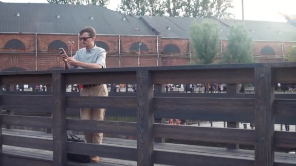 Joven usando un smartphone parado en un puente de madera. Parque de la ciudad al fondo . — Vídeo de stock