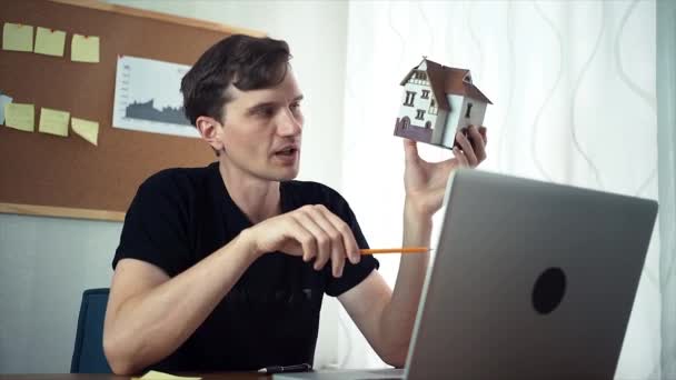 Молодой дизайнер с видеозвонком с помощью ноутбука демонстрирует модель дома — стоковое видео