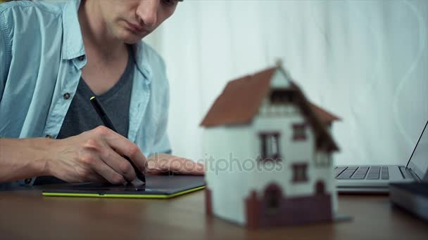 Diseñador usando tableta gráfica creando proyecto de vivienda. Modelo de casa sobre la mesa — Vídeos de Stock