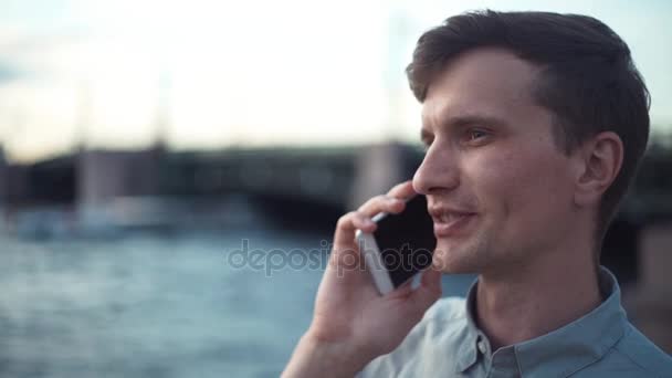 若い男は、屋外で立って電話で話しています。背景の橋. — ストック動画
