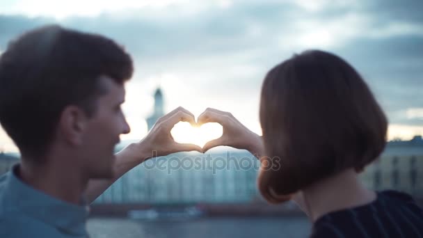 Feliz joven pareja de pie al atardecer haciendo una forma de corazón con sus manos — Vídeos de Stock