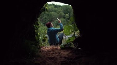 Mağara çıkış yakınında oturan smartphone kullanan adam. Arka plan, derin orman.