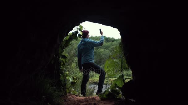 Ο τύπος χρησιμοποιώντας smartphone στέκεται κοντά σε έξοδο από το σπήλαιο. Πυκνό δάσος στο παρασκήνιο. — Αρχείο Βίντεο