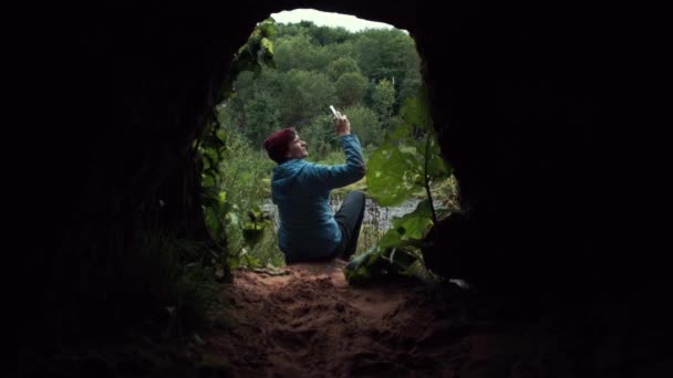 Homem usando smartphone sentado perto da saída da caverna. Floresta profunda no fundo . — Vídeo de Stock