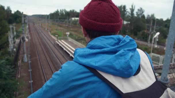 Πεζοπόρος ντυμένος με σακάκι και πλεκτά καπέλο με σακίδιο στέκεται στην σιδηροδρομική γέφυρα — Αρχείο Βίντεο