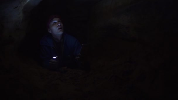 Путешественник с помощью смартфона лежит на земле в тесном отверстии в пещере с фонариком — стоковое видео