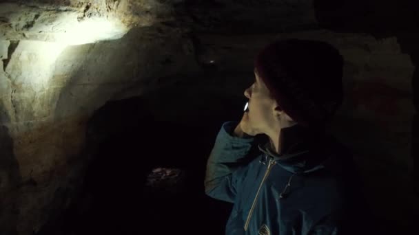 Young guy standing in tight cave explores walls with help flashlight in his hand — Stock Video