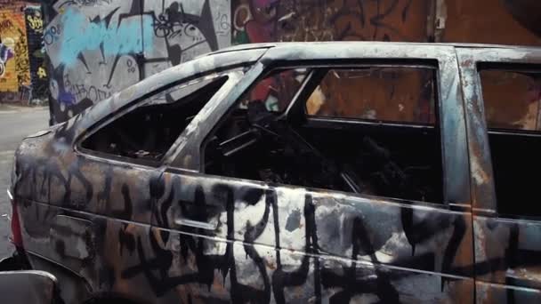 Chocó coche quemado de pie al aire libre. Grafitis no identificables en las paredes alrededor — Vídeo de stock