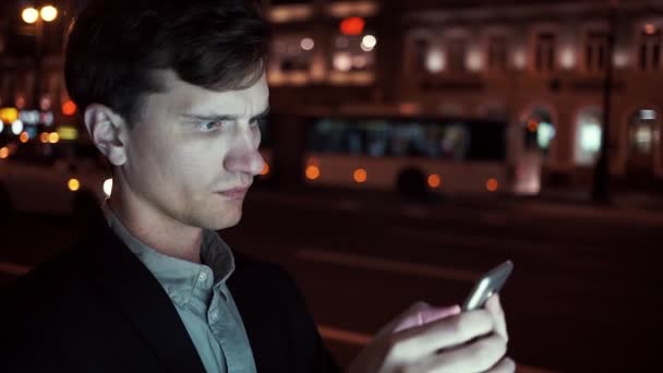 Junger Mann surft nachts mit seinem Handy auf einer belebten Stadtstraße — Stockvideo