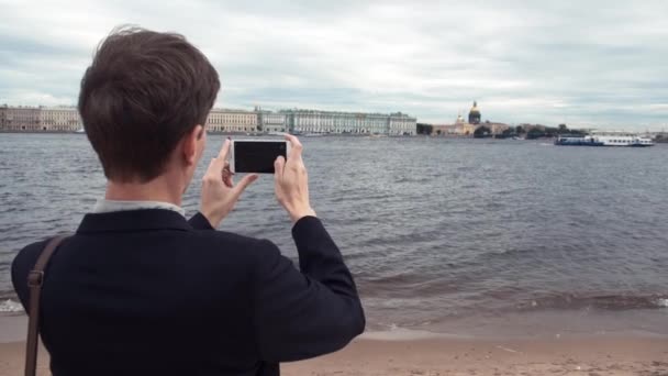 Молодий чоловік фотографує за допомогою смартфона, що стоїть на набережній міста — стокове відео